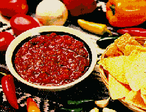 A bowl of salsa with tortilla chips in a basket, surrounded by tomatoes, peppers, and an onion.